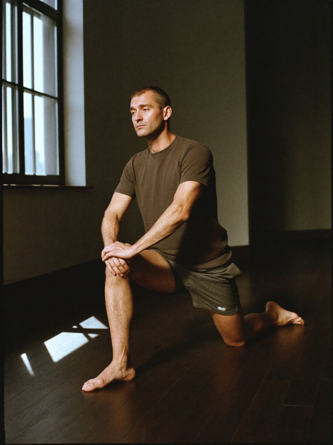 A man part way through a slow half kneeling stretch on a dark wooden floor.